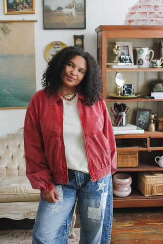 Woman wearing a red vintage washed bomber jacket over a white top, paired with patchwork jeans, standing in a vintage-style room.