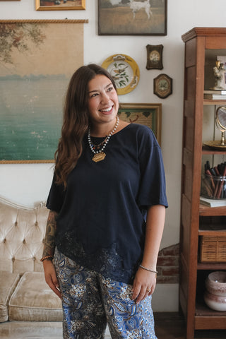 A woman smiling in a cozy room, wearing a dark navy blue short-sleeve top with lace detailing at the hem and blue paisley patterned pants.