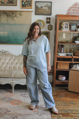 Woman standing in a living room with vintage decor wearing a light blue lace lined tee and light wash barrel jeans
