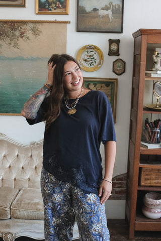 A woman stands in a decorated room wearing a navy blue short-sleeve top and blue paisley patterned pants. The background features a tufted sofa, a wooden cabinet, and various framed wall art pieces.