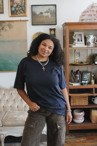 A woman with curly black hair stands in a cozy, decorated room wearing a navy blue short-sleeve t-shirt with a lace hem and brown cargo pants.