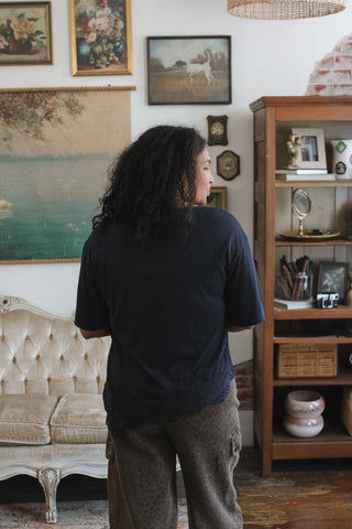 A person with curly dark hair stands with their back to the camera in a cozy, vintage-style room, wearing a dark navy blue t-shirt and brown patterned pants. The room features a tufted cream sofa, a wooden bookshelf, and framed art on the walls.
