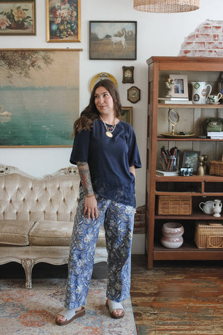 A woman stands in a vintage-style room wearing a navy blue short-sleeve top and blue floral wide-leg pants. The room features a tufted cream sofa, a wooden bookshelf, and framed wall art.