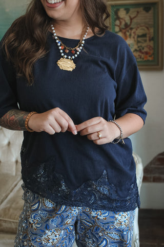 A woman wearing a navy blue top with a lace hem and blue paisley patterned pants, accessorized with layered necklaces and bracelets.