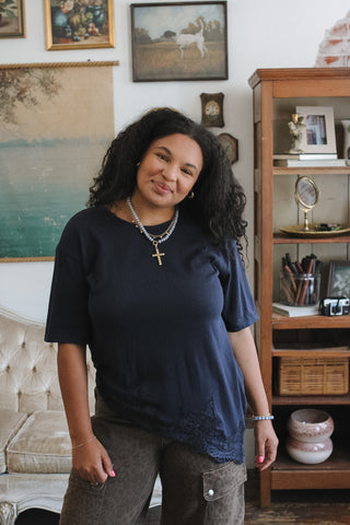 A woman with curly black hair stands in a decorated room wearing a dark navy blue short-sleeve t-shirt and brown cargo pants. She is wearing layered silver necklaces, one with a gold cross.
