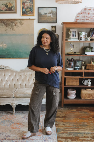 A woman stands in a styled room wearing a dark navy blue short-sleeve top and brown wide-leg cargo pants. The background features a vintage tufted sofa, a wooden bookshelf, framed wall art, and a rustic wood floor.