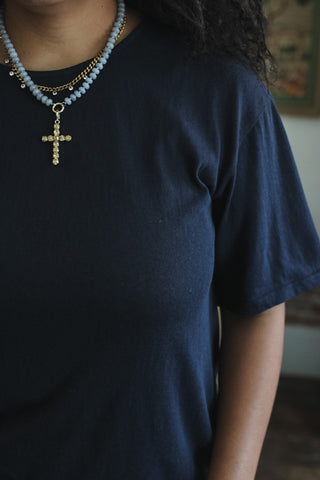 A close-up of a person wearing a dark navy blue t-shirt and a layered necklace featuring light blue beads, a gold chain, and a gold cross pendant.