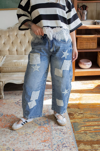 Person wearing a striped shirt and patchwork jeans in a room with a couch and shelves.