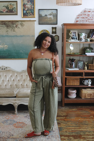 A woman stands in a decorated room wearing a sage green strapless jumpsuit with barrel legs and a drawstring waist, paired with red sandals and a patterned shoulder bag.