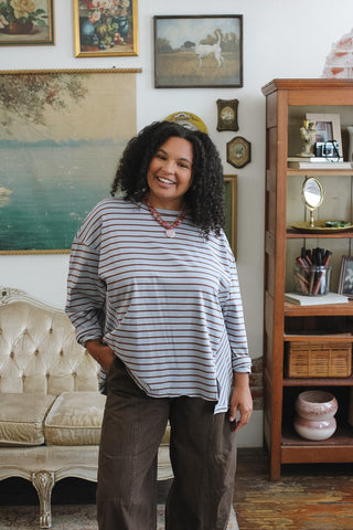 A woman with curly black hair stands in a vintage-style room wearing a blue and brown striped long-sleeve top and brown pants. The background features framed artwork on a white wall, a cream tufted sofa, and a wooden cabinet.