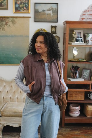 A woman with curly black hair stands in a vintage-style room wearing a brown vintage washed bomber zip up vest over a blue and brown striped long-sleeve shirt and light blue jeans. She is holding a brown shoulder bag, with a tufted sofa, framed paintings, and a wooden bookshelf in the background.