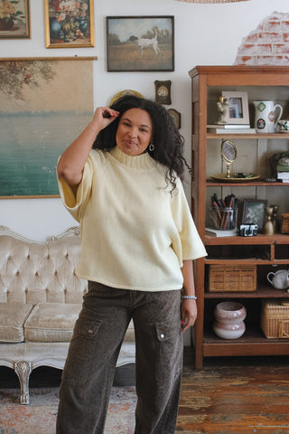 A woman wearing a cream-colored short-sleeve mock neck knit sweater and dark brown wide-leg cheetah pants, standing in a vintage-style room with a wooden bookshelf and framed art.