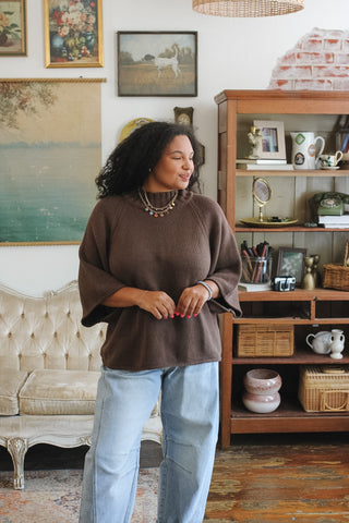 A woman wearing a brown knit top with wide bell sleeves and light blue jeans, standing in a cozy, vintage-style room with a wooden bookshelf and framed art.