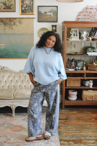 A woman stands in a styled room wearing a light blue knit mock neck sweater and blue patterned wide-leg pants, with sandals, in front of a wooden bookshelf and vintage sofa.