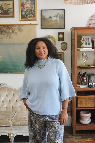 A woman stands in a decorated room wearing a light blue ribbed mock-neck tunic top with wide bell sleeves and patterned blue, gray, and beige pants.