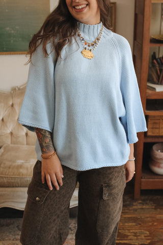 A woman wearing a light blue ribbed knit mock-neck sweater with wide three-quarter sleeves, paired with brown cargo pants, in a cozy indoor setting.
