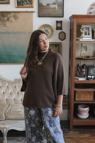A woman wearing a brown knit mock neck sweater top and blue floral wide-leg pants, standing in a decorated room with vintage furniture and framed art.