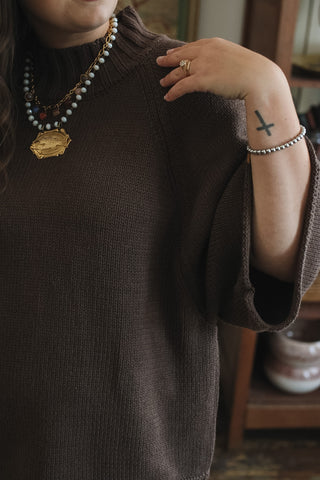A close-up of a person wearing a dark brown knit mock neck sweater, layered with multiple necklaces including a gold pendant and a pearl strand, along with bracelets and rings.