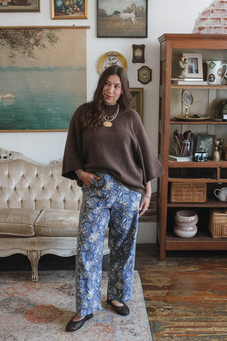 A woman stands in a vintage-style room wearing a brown knit mock neck sweater and blue floral wide-leg pants. The room features a tufted sofa, a wooden bookshelf, and framed wall art.