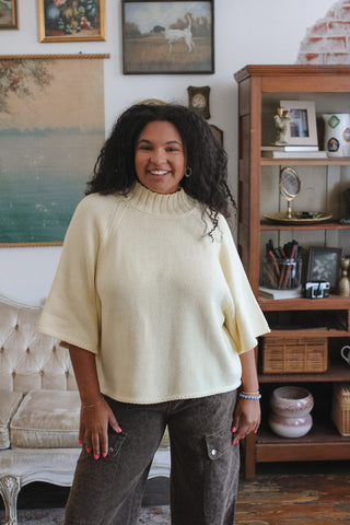 A woman with curly black hair stands in a cozy, vintage-style room wearing a cream-colored knit mock-neck sweater and dark gray cargo pants.