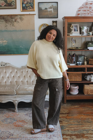 A woman stands in a vintage-style room wearing a pale yellow knit mock-neck tunic sweater and dark brown wide-leg corduroy pants. The room features a tufted sofa, a wooden bookshelf, and framed art on the walls.