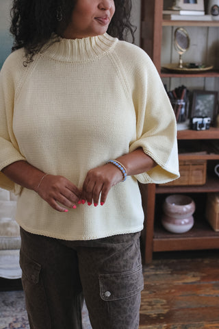 A woman wearing a cream-colored knit mock-neck sweater with elbow-length flared sleeves, paired with dark brown cargo pants, standing in a home interior setting.