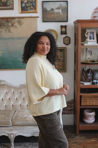 A woman with curly black hair stands in a vintage-style room wearing a cream-colored knit mock-neck top and dark brown pants. The background features a tufted sofa, a wooden bookshelf, and framed wall art.