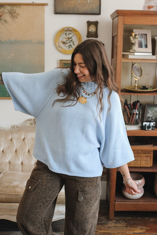 A woman wearing a light blue knit sweater with wide, draped sleeves and brown cheetah pants, standing in a cozy, vintage-style room with a wooden cabinet and framed art.