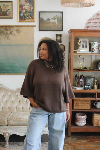 A woman wearing a brown knit mock neck sweater and light blue jeans stands in a decorated room with framed art, a tufted sofa, and a wooden shelving unit.