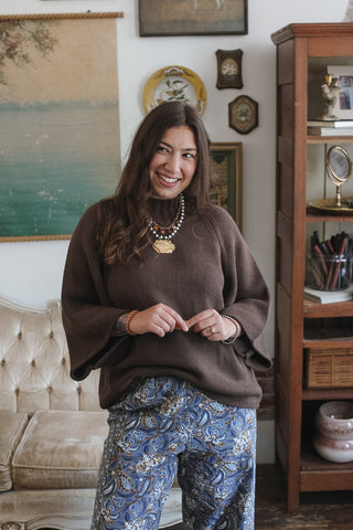 A woman smiling in a cozy indoor setting, wearing a brown mock neck knit sweater with wide bell sleeves and blue paisley pants. She is accessorized with layered gold necklaces and bracelets.