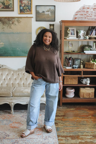 A woman stands in a styled room wearing a brown mock neck knit sweater, light blue jeans, and white slide sandals. The room features a wooden bookshelf, a tufted sofa, and framed artwork on the wall.
