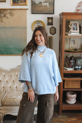 A woman wearing a light blue ribbed knit mock neck sweater and brown cargo pants, standing in a vintage-style room with a wooden bookshelf and framed art.