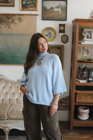 A woman wearing a light blue knit mock neck poncho sweater and brown pants, standing in a vintage-style room with a wooden bookshelf, tufted sofa, and framed wall art.