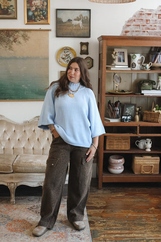 A woman stands in a vintage-style living room wearing a light blue oversized mock neck knit sweater with wide bell sleeves and dark brown wide-leg trousers.