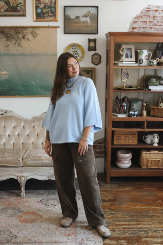A woman stands in a vintage-style room wearing a light blue mock neck knit sweater brown wide-leg trousers, and beige shoes. The room features a tufted sofa, a wooden bookshelf, and framed art on the wall.