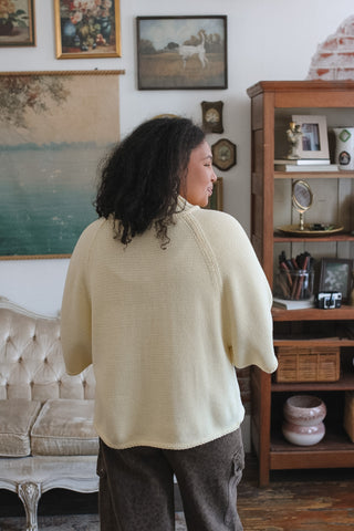 A person with curly dark hair stands with their back to the camera wearing a cream-colored knit mock neck sweater over dark brown pants in a cozy, vintage-style room with framed art and a wooden cabinet.