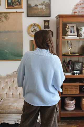 A woman viewed from behind wearing a light blue knit mock neck sweater and brown pants, standing in a vintage-style room with a wooden cabinet, framed art, and a tufted sofa.