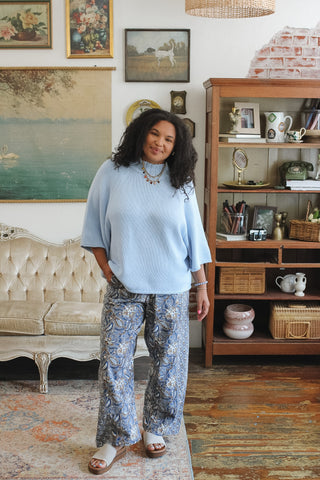 A woman stands in a styled room wearing a light blue ribbed knit mock-neck knit sweater and blue-and-white patterned wide-leg pants, paired with brown slide sandals.