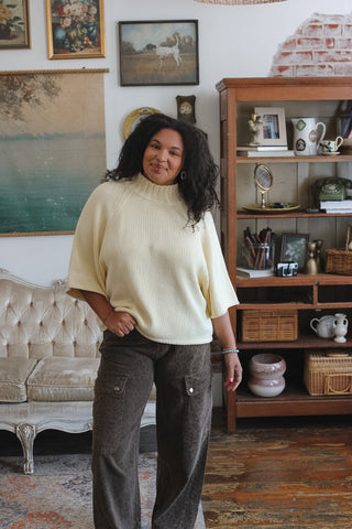 A woman wearing a cream ribbed mock-neck short-sleeve top and dark brown cheetah wide-leg cargo pants, standing in a cozy, vintage-style room with a tufted sofa and a wooden bookshelf.