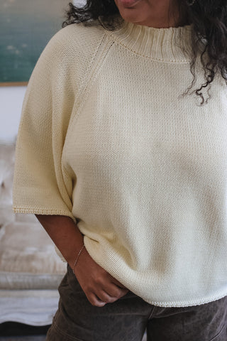 A person wearing a cream-colored knit short-sleeve sweater with a mock neck, paired with dark brown pants, shown in a close-up lifestyle shot.