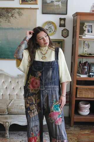 A woman wearing a cream t-shirt and dark blue denim overall with colorful floral and graphic patches, standing in a vintage-style room with a tufted sofa and wooden shelving.