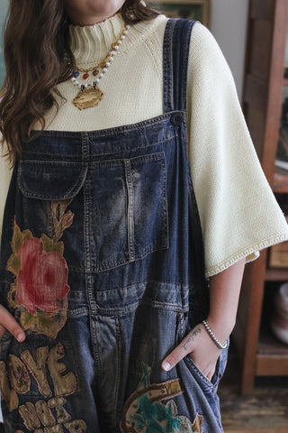 A person wearing dark blue denim overalls with a large red rose graphic and distressed patches, layered over a cream-colored knit turtleneck sweater.