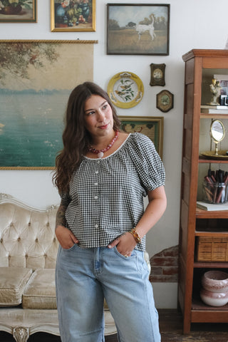 A woman wearing a black and white gingham puff-sleeve top and light blue jeans, standing in a vintage-style room with framed art, a tufted sofa, and a wooden cabinet.