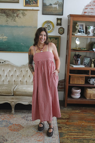A woman wearing a red and white gingham maxi dress with spaghetti straps and side pockets, standing in a vintage-style room with a tufted sofa and a wooden bookshelf.