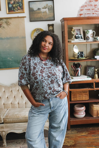 Woman wearing a blue floral puff-sleeve blouse and light denim jeans in a cozy living room.