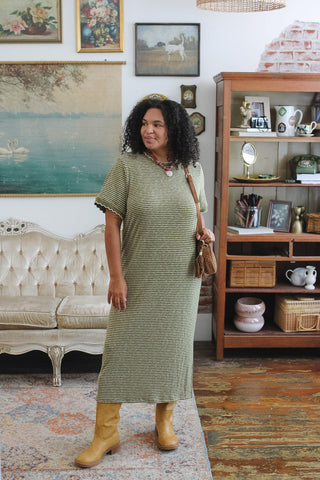 A woman wearing a ribbed knit striped olive green midi dress with short sleeves and tan boots, standing in a vintage-style room with a tufted sofa and wooden shelving.