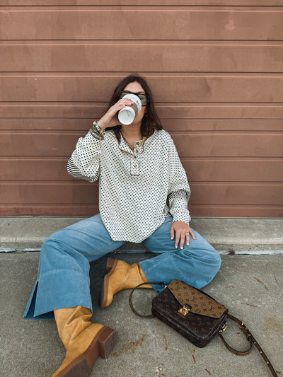 Girl sitting on the ground with a coffee cup, wearing a white and black polka dot shirt, blue jeans, and brown boots, against a brown wall.