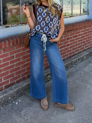 Woman wearing the Bennet medium wash high rise wide leg jeans with a white drawstring waist, styled with a brown and blue floral knit vest and suede loafers