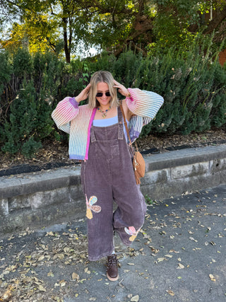 Woman wearing the Tate patchwork corduroy overalls in washed mocha with flower patchwork on the legs, styled with a rainbow striped cardigan and lace-up boots