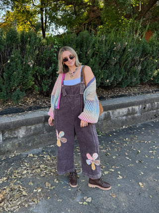 Woman wearing the Tate patchwork corduroy overalls in washed mocha with flower patchwork on the legs, styled with a rainbow striped cardigan and lace-up boots